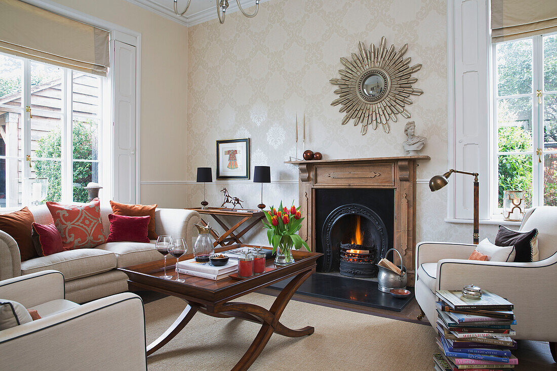 Wooden coffee table and stack of books with sunburst mirror above lit fire in Worcestershire home England UK