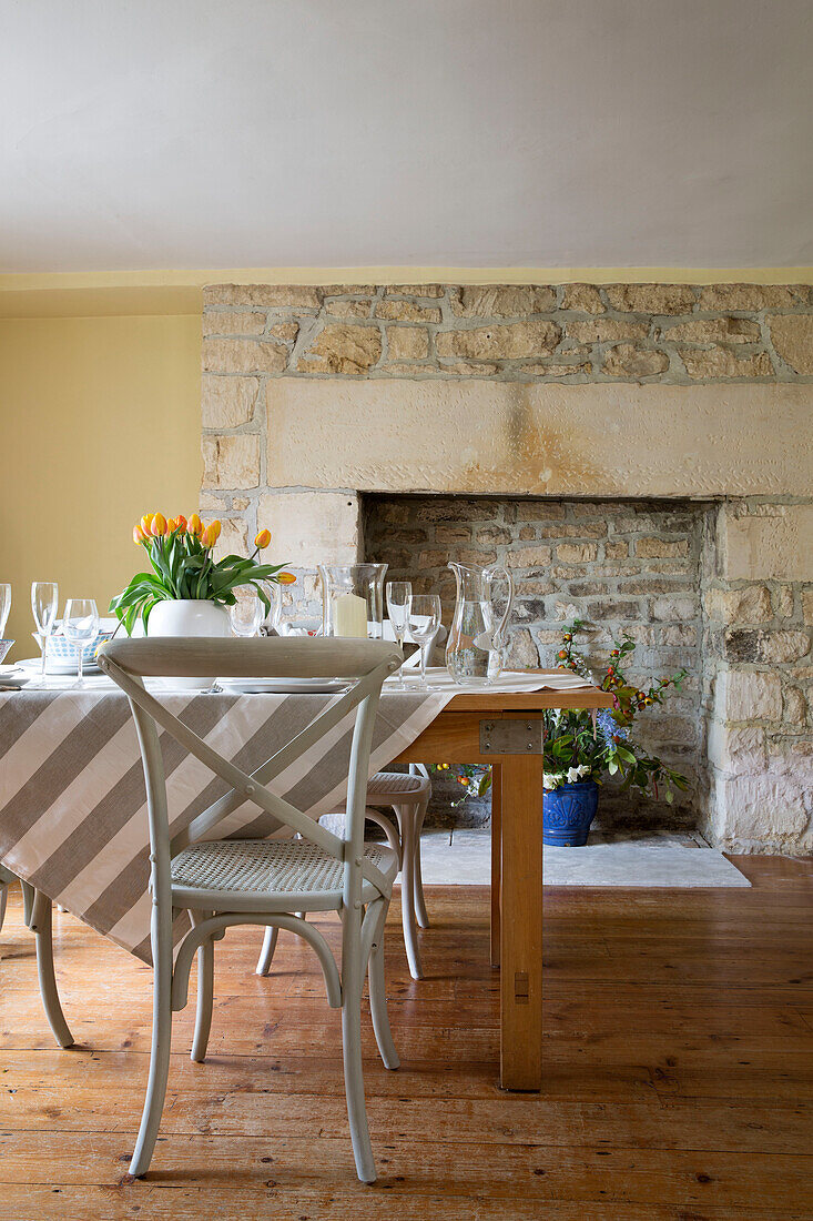 Bemalter Stuhl und gestreifte Tischdecke mit offenem Steinkamin in einem Bauernhaus in Gloucestershire, England, UK