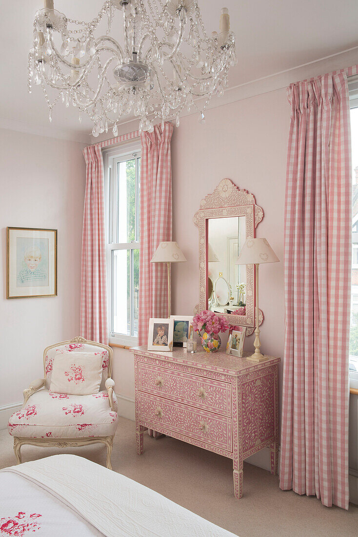 Pink gingham curtains and decorative chest of drawers with floral chair and chandelier in bedroom of Victorian terrace house South London UK