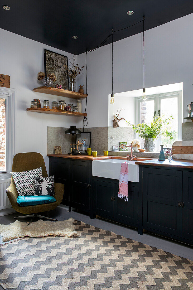 Retro-Stühle in der Küche mit schwarz gestrichenen Schränken und Butler-Spüle am Fenster in einer umgebauten walisischen Scheune, UK