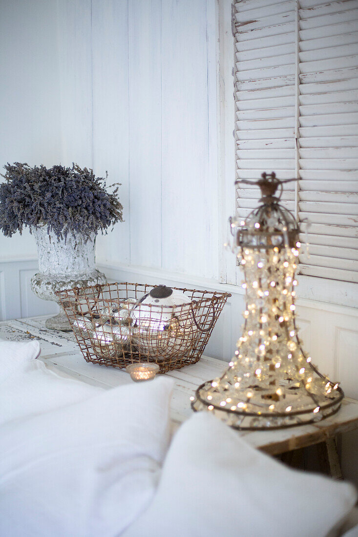 Scandi style living room detail with white cushions lavender in a vintage planter and galvanised basket filled with silver baubles a chandelier wrapped with fairylights and louvered shutters on the wall