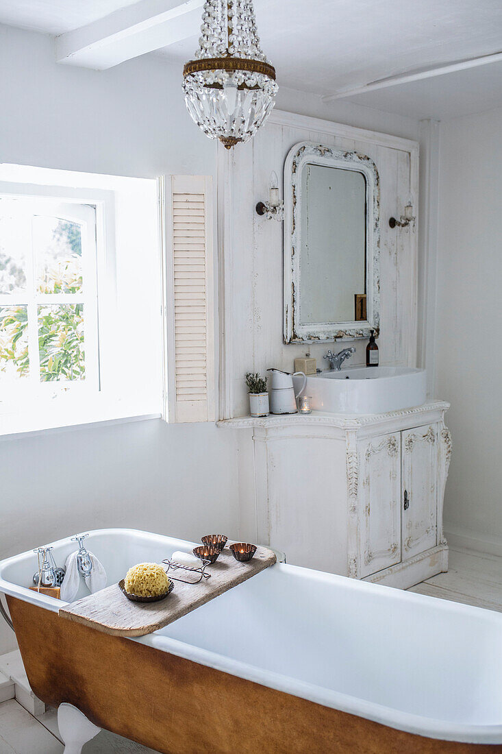 White Scandinavian style bathroom with ornate carved vanity unit bath beside the window and white enamelware and pretty crystal chandelier