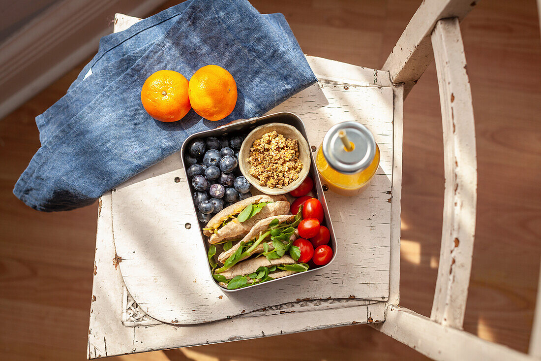 Lunchbox mit gefülltes Pitabrot, Granola, Tomaten und Blaubeeren auf einem Stuhl