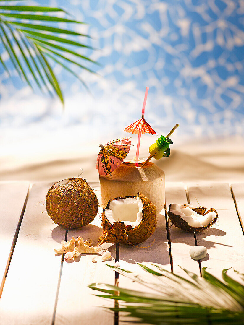 Coconut, one broken, one whole, and a wooden cocktail cup against a tropical backdrop