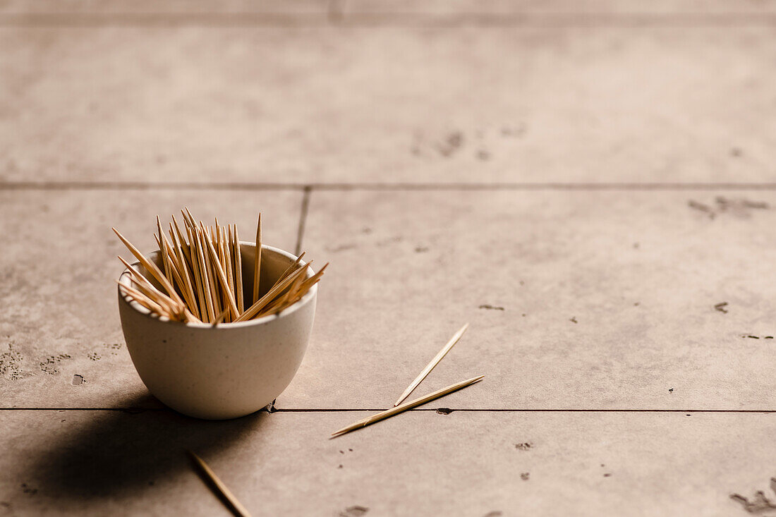 A small bowl of toothpicks on the edge of a table with a few scattered on the table