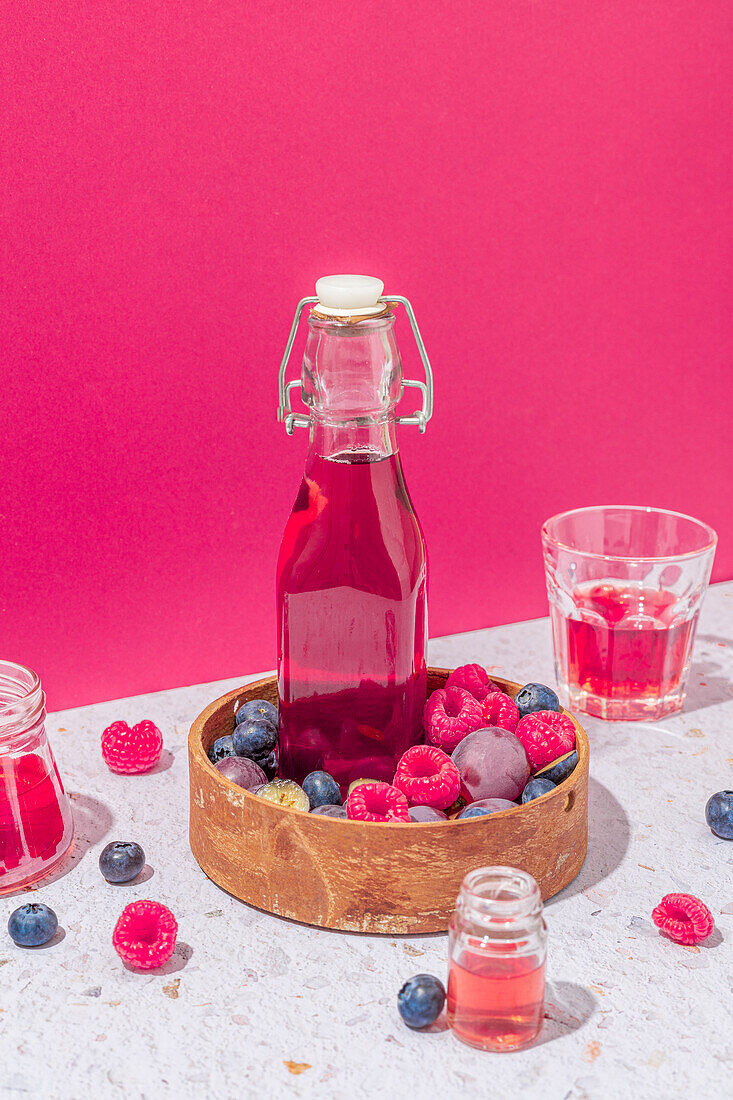Glass bottle of fresh fruits juice in wooden bowl with ripe berries served on table with glasses on pink background