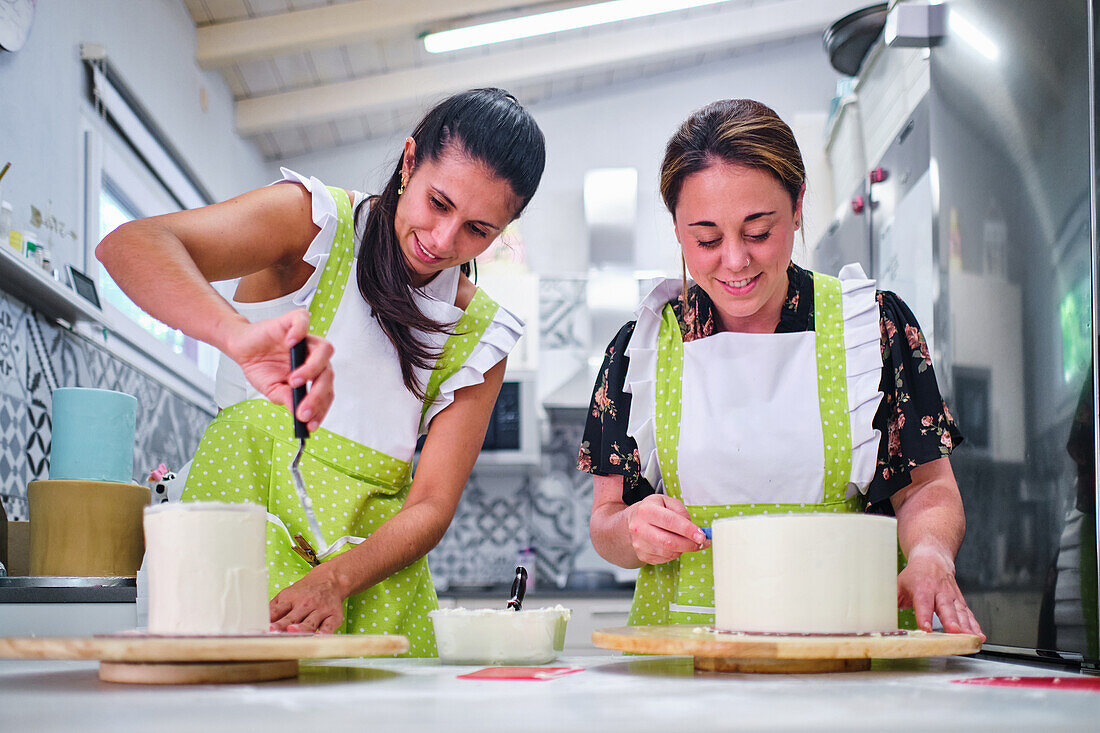 Concentrated confectioners in aprons standing and spreading cream on sponge cakes in light kitchen