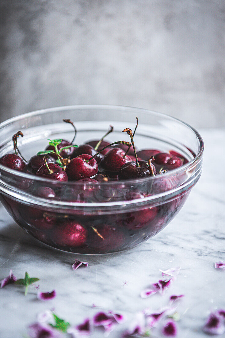 Glass bowl full of fresh ripe cherries placed on marble table with tender petals