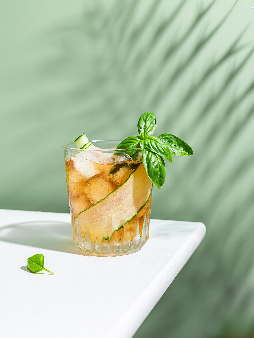 Cocktail mit Gurkenscheibe und Basilikumblättern vor grünem Hintergrund mit Schattenwurf