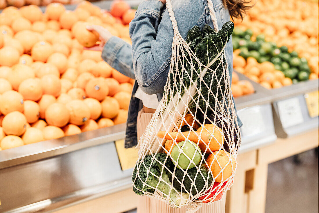 Junge Frau mit Gemüse und Obst in umweltfreundlicher Netztasche steht vor Obstregal im Supermarkt