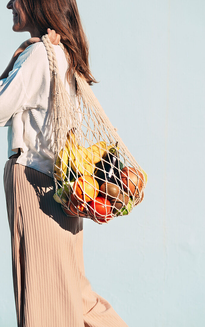 Side view of cropped smiling female standing with assorted fruits and vegetables in eco friendly mesh bag against blue wall
