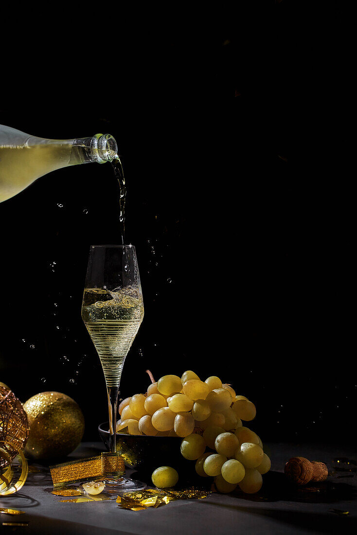 Champagne pouring from bottle into wineglass placed on table with fresh ripe grapes during Christmas celebration