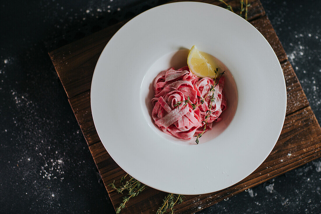Vegan pink beetroot pasta with lemon slice and thyme sprig served on wooden board on black table