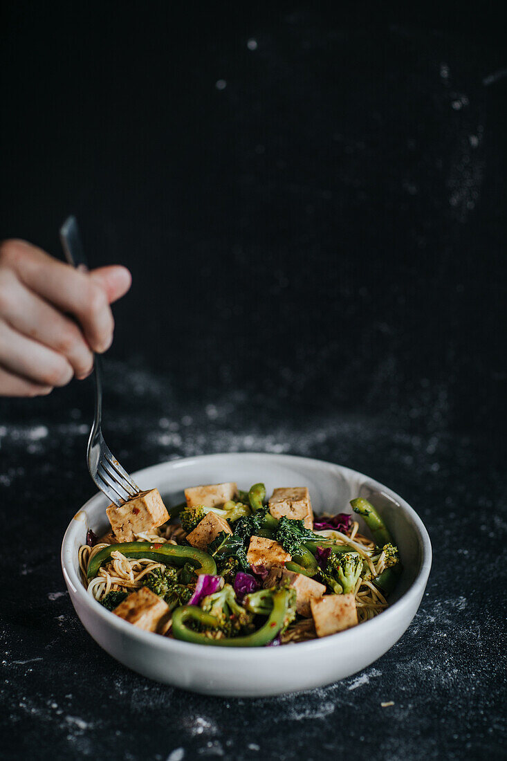 Crop unrecognizable person eating appetizing vegetarian salad with fried tofu and vegetables served on black background