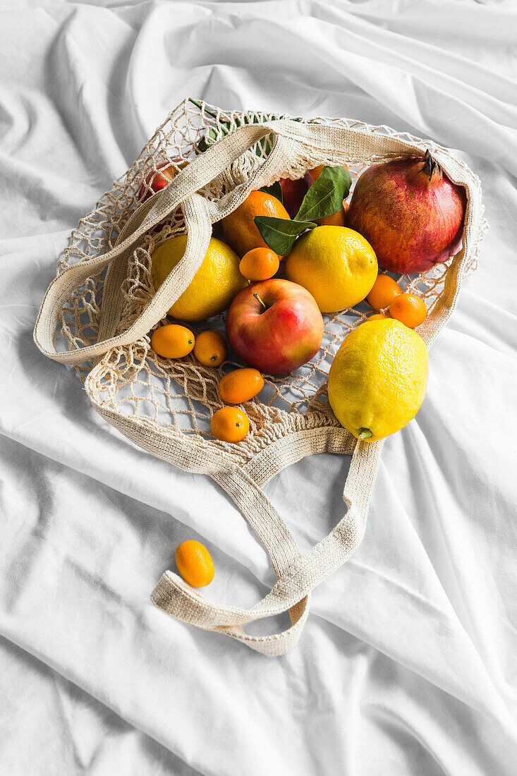 Different whole ripe apple with lemons near pomegranate and kumquats in eco bag on creased cloth