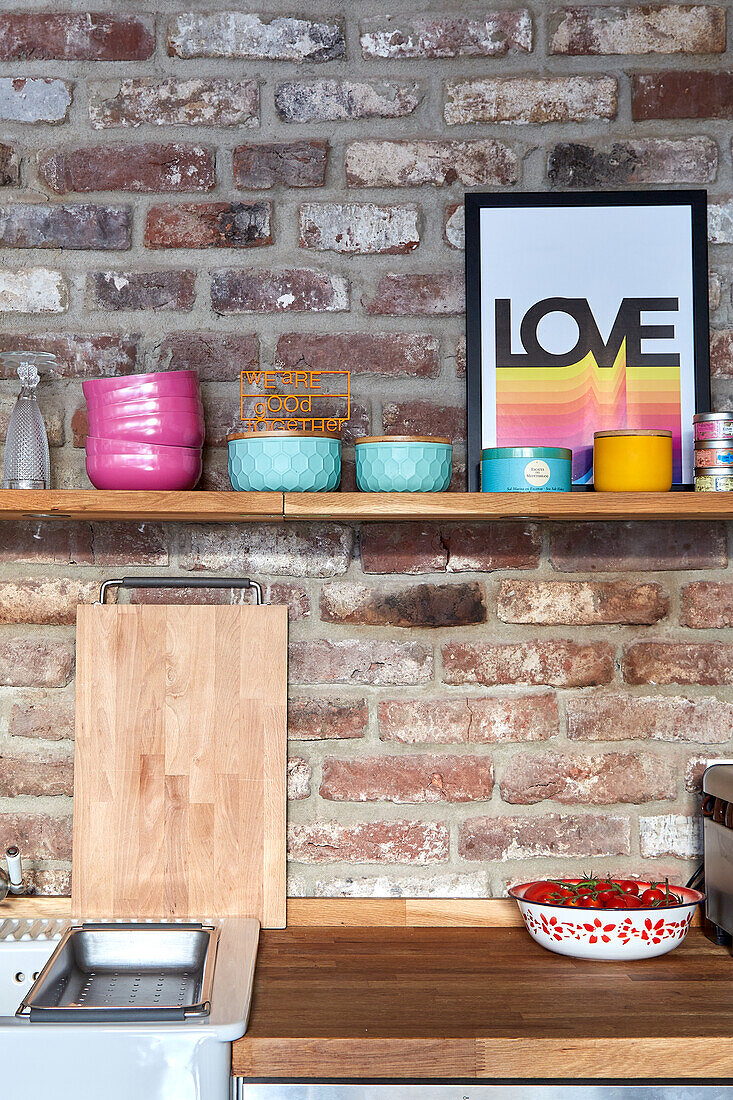 Wooden shelf with colourful bowls above kitchen worktop on brick wall