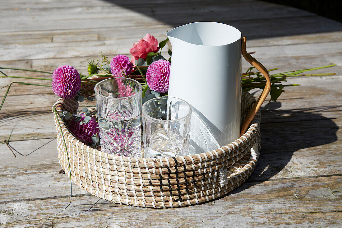 Woven tray with pitcher, glasses and dahlias on a wooden background