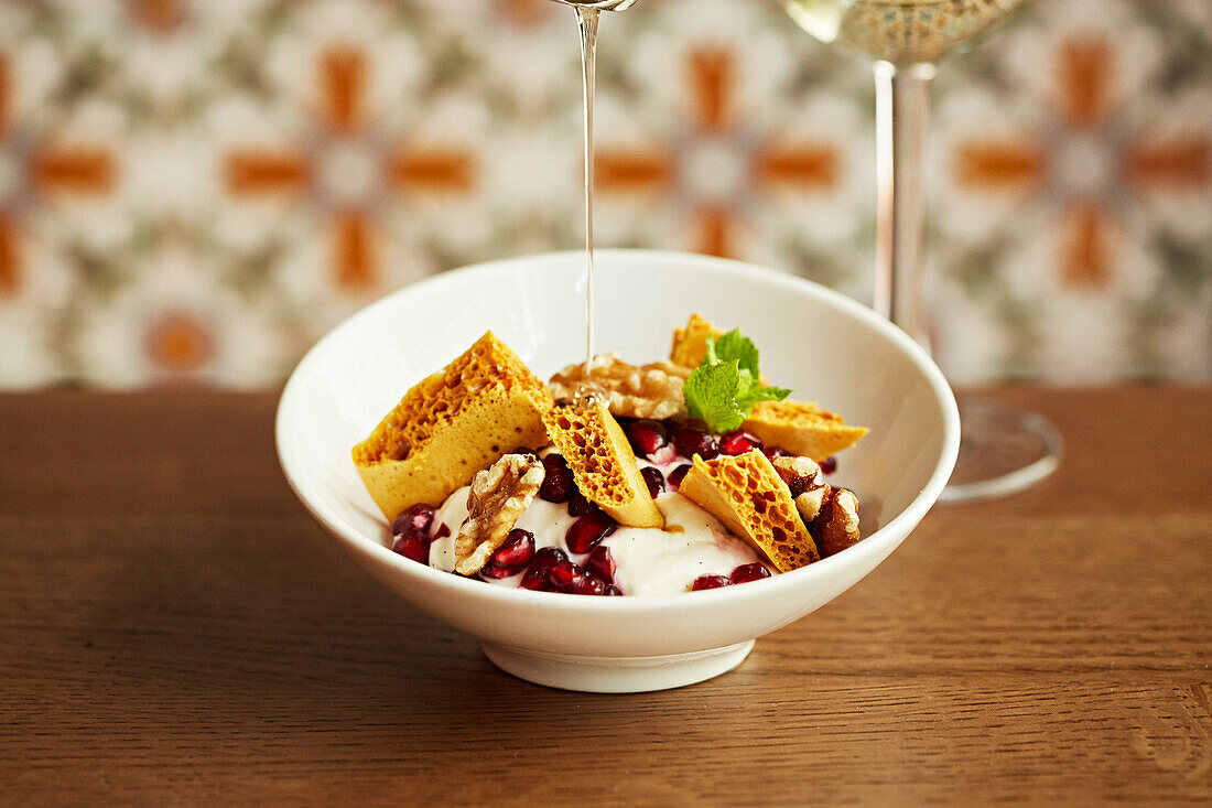 Brittle honeycomb pieces in yoghurt with pomegranate and walnut with honey being poured on top