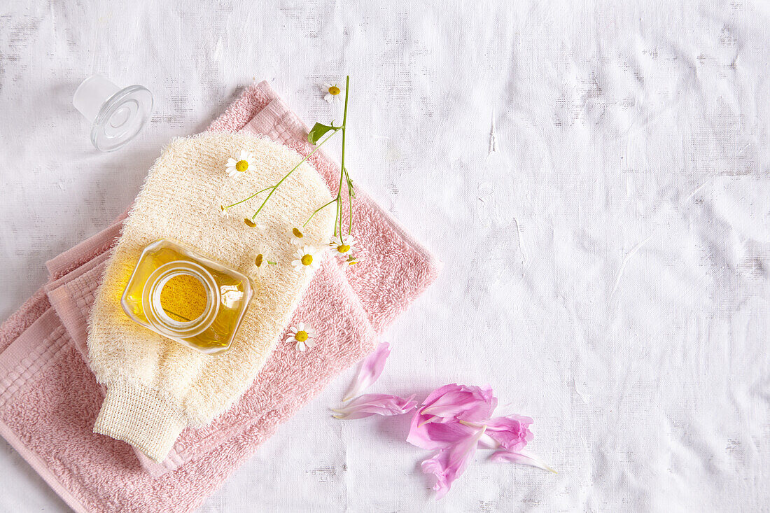 Ölfläschchen auf einem Massagehandschuh und Handtüchern, römische Kamilleblüten und Rosenblüten