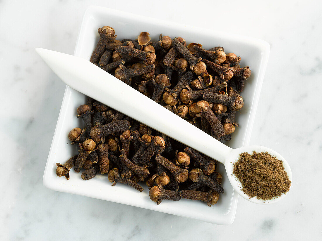 A bowl with cloves and a spoon with clove powder on a light background