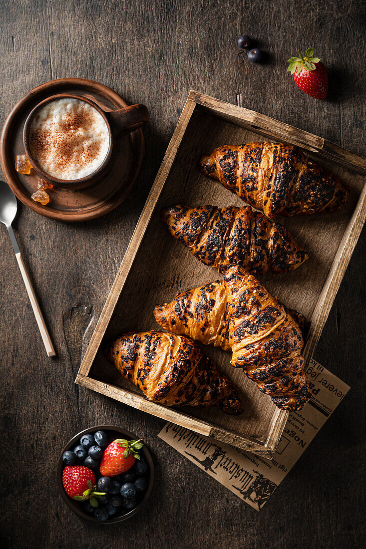 Croissants in a wooden tray with a cup of coffee and fresh berries on a wooden table