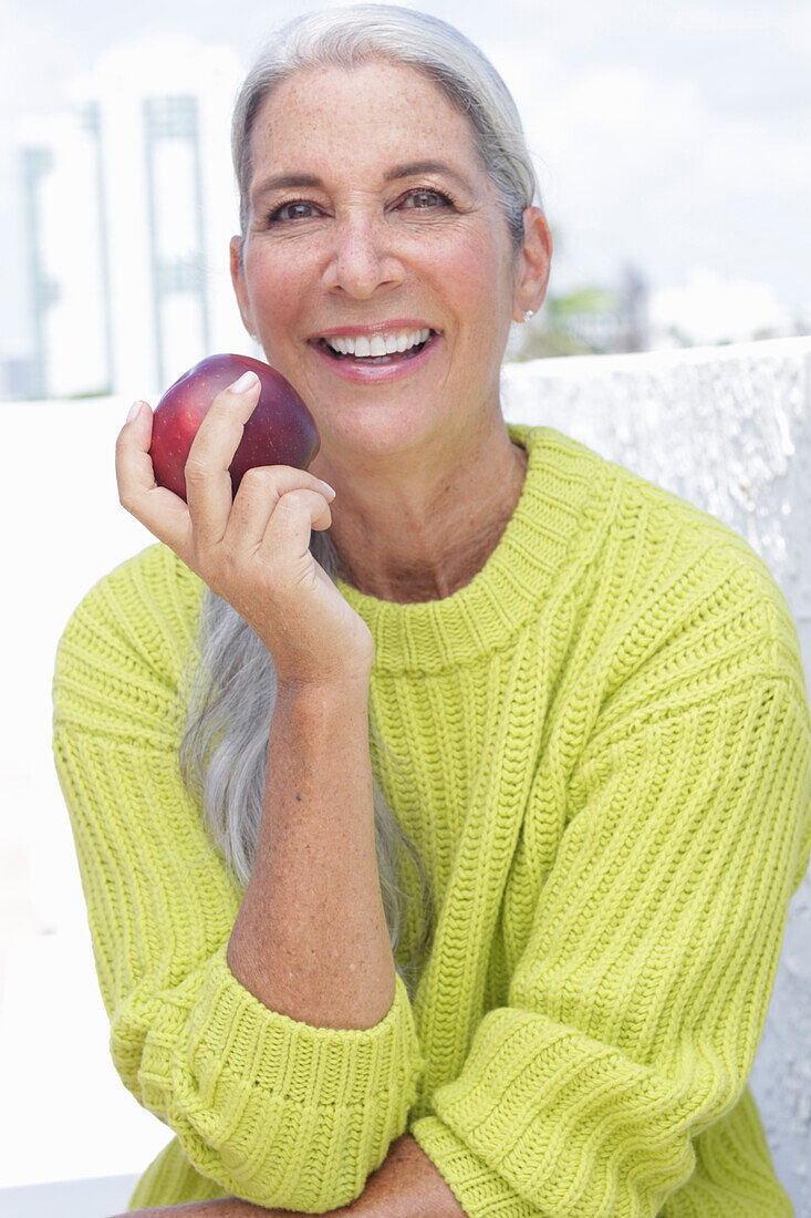 A grey-haired woman wearing a greenish-yellow knitted jumper holding an apple