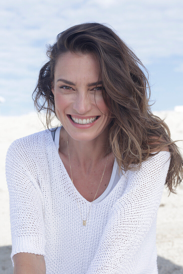 A happy, long-haired woman on a beach wearing a white jumper