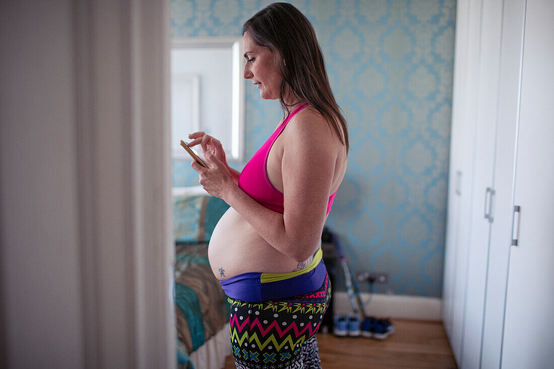 Pregnant woman in sports bra using smart phone in doorway