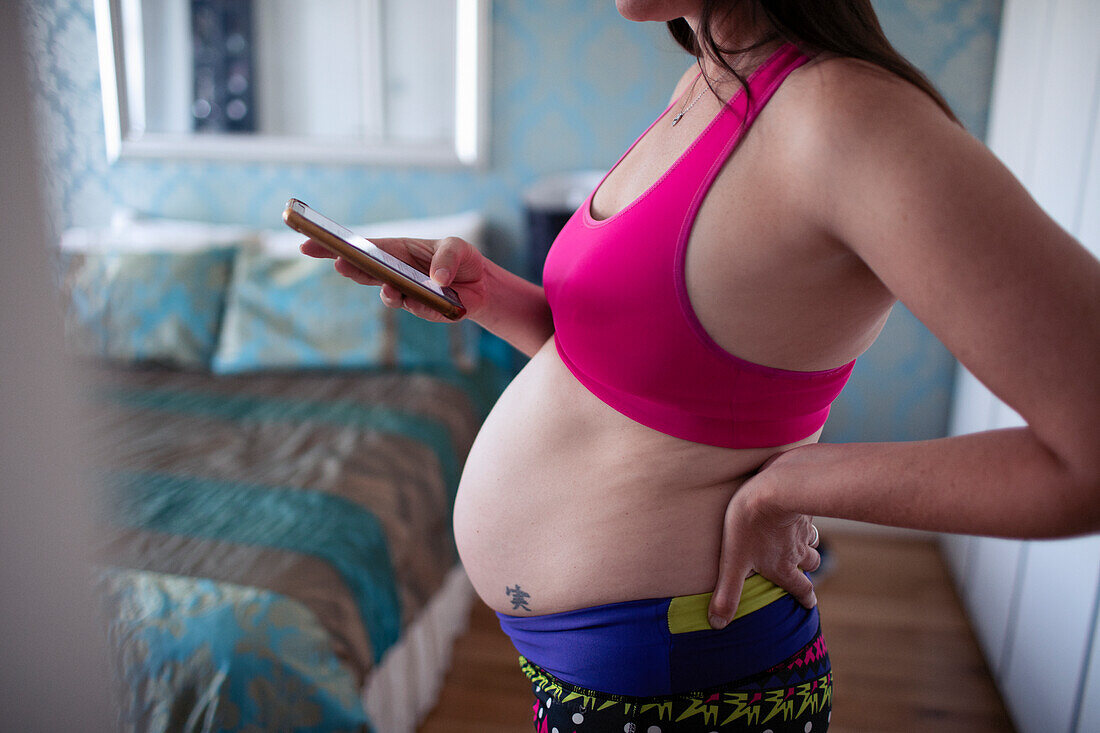 Pregnant woman in sports bra using smart phone in bedroom