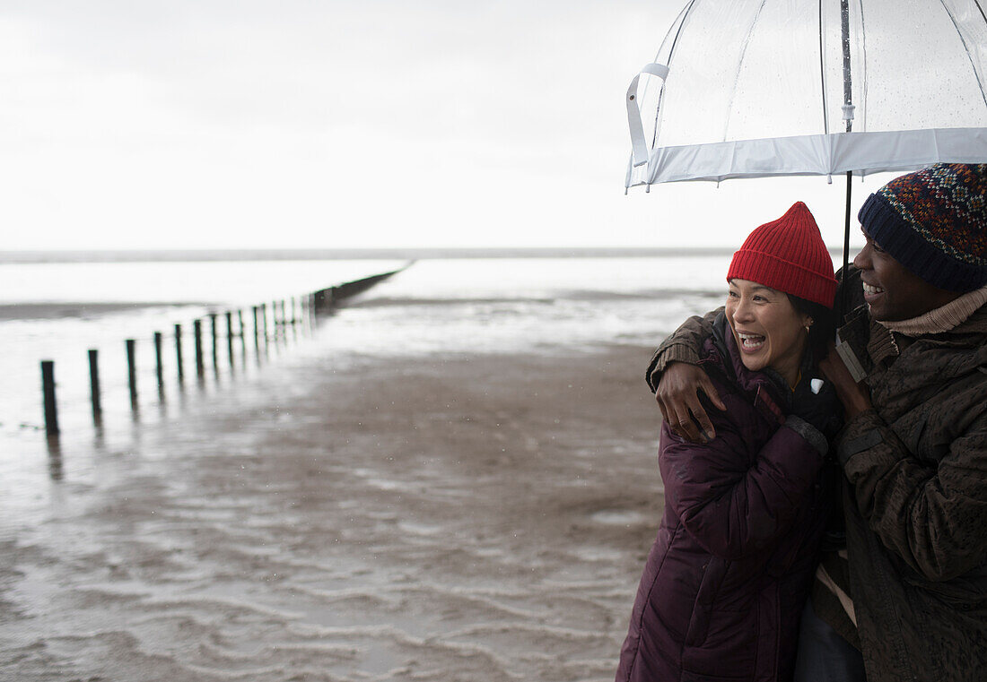 Happy couple hugging under umbrella on wet winter beach