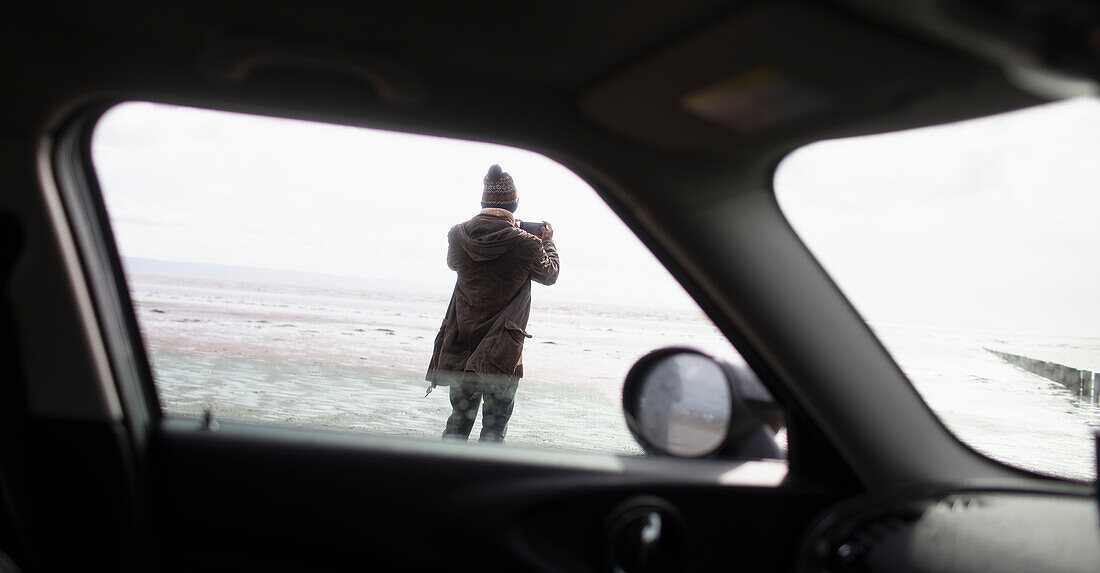 Man with camera phone on winter beach outside car