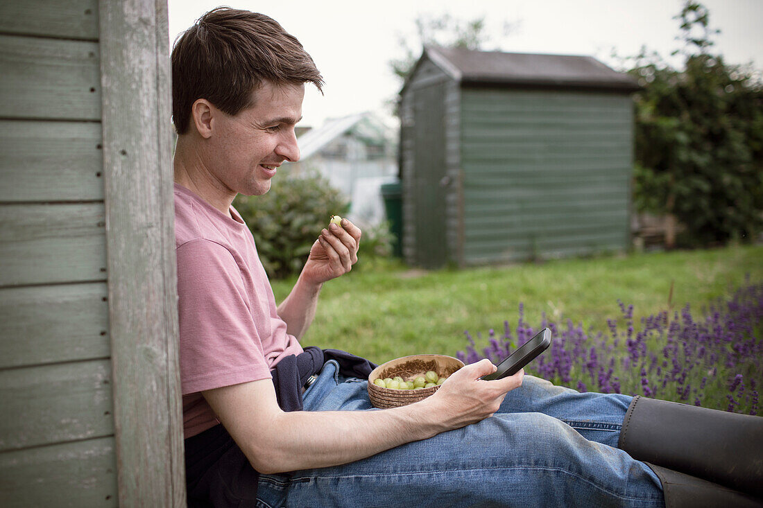 Smiling man eating gooseberries and using smart phone in garden