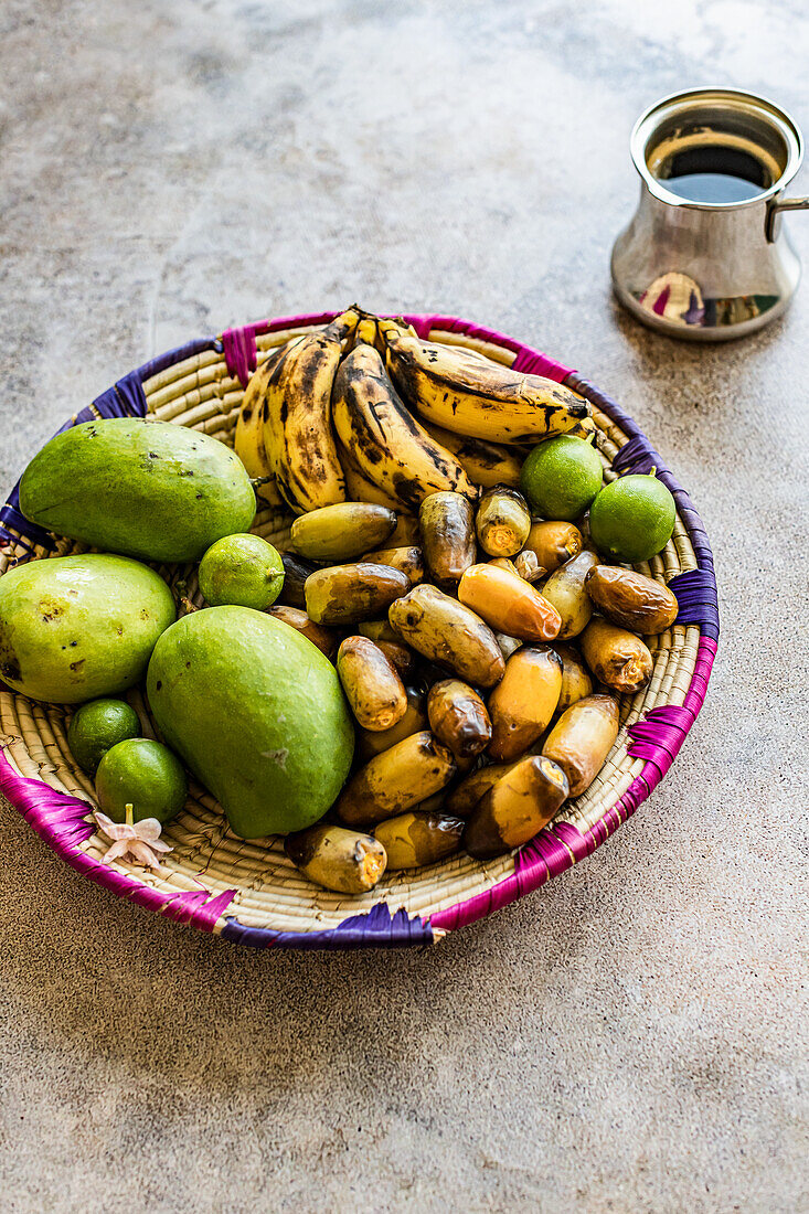 Fruit basket with semi-ripe dates, bananas, and green mango, next to Arabic coffee