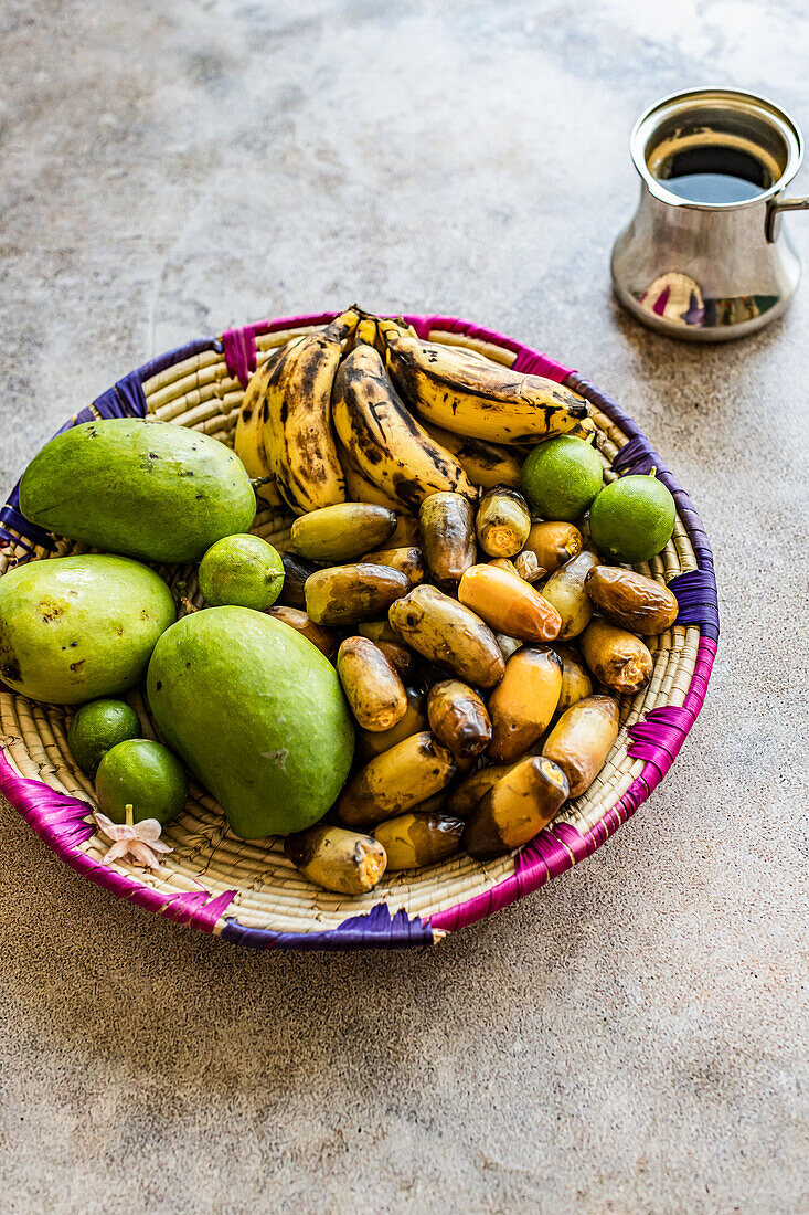 Obstkorb mit halbreifen Datteln, Bananen und grünen Mango, daneben arabischer Kaffee