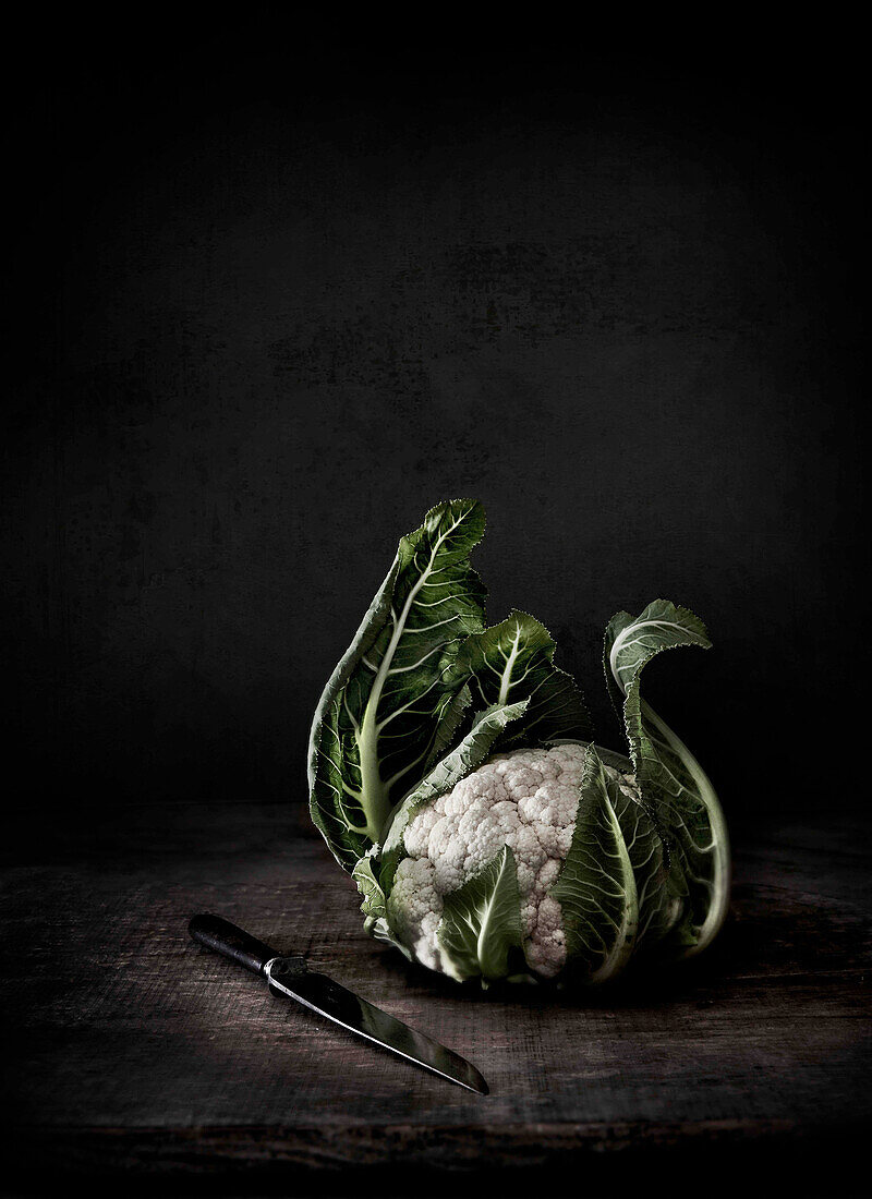 Cauliflower with knife on wooden table in front of black background