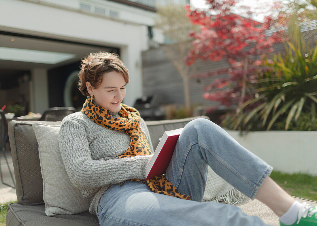 Young woman reading book on sunny patio lounge chair
