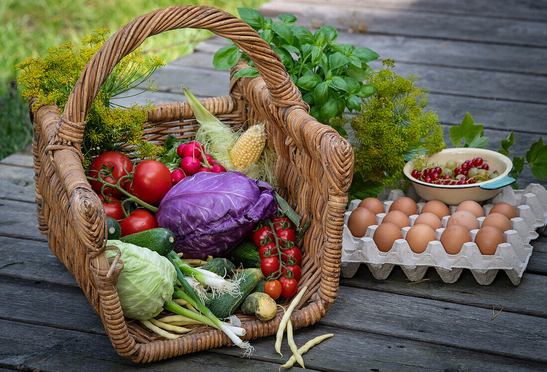 Frisches Gemüse im Korb daneben Kräuter, Eierpalette und Schälchen mit Beeren