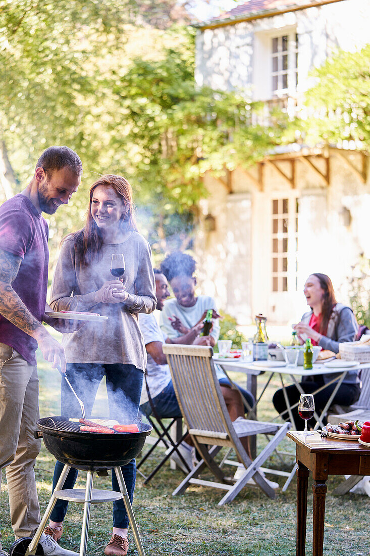 Young man preparing food in barbecue grill while his friends having drinks in background