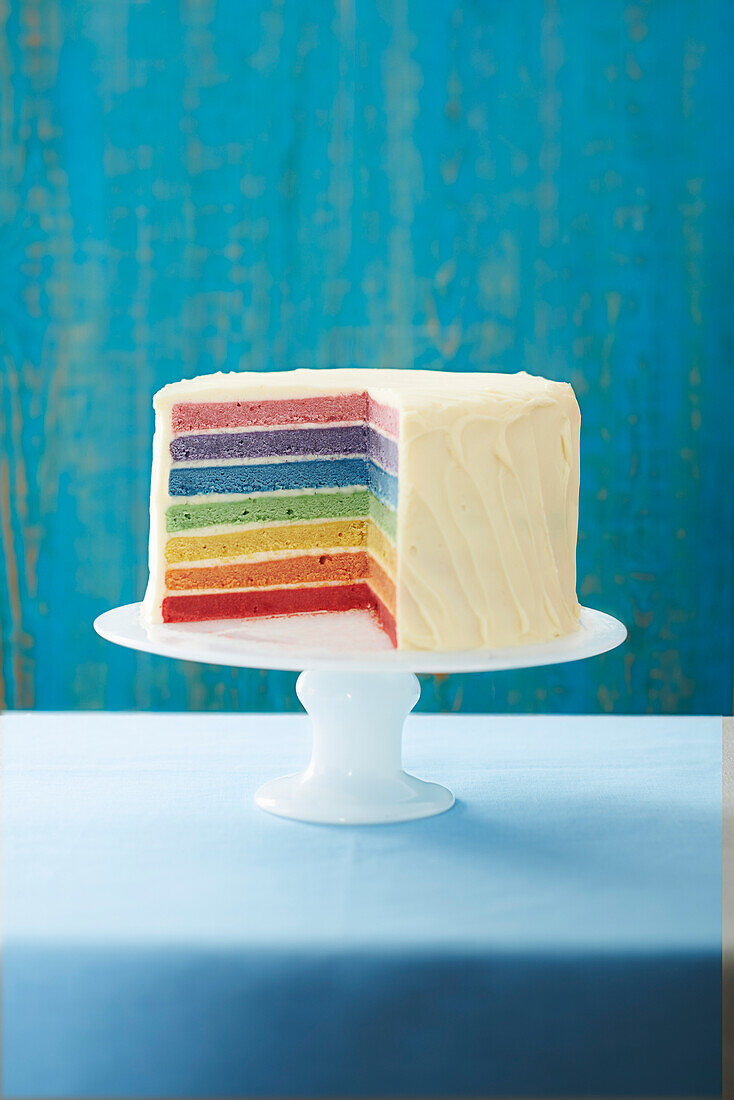 Regenbogentorte, angeschnitten auf Tortenständer vor blauem Hintergrund