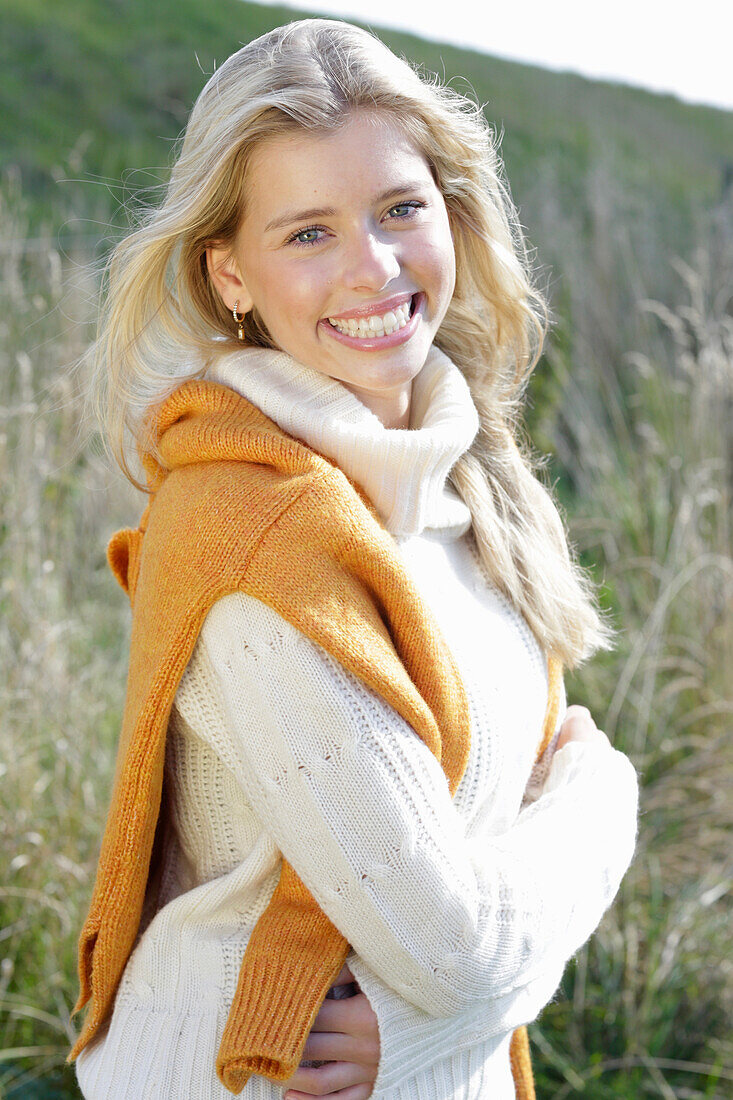 Young blond woman in white turtleneck sweater, with yellow knitted sweater slung over shoulders in nature