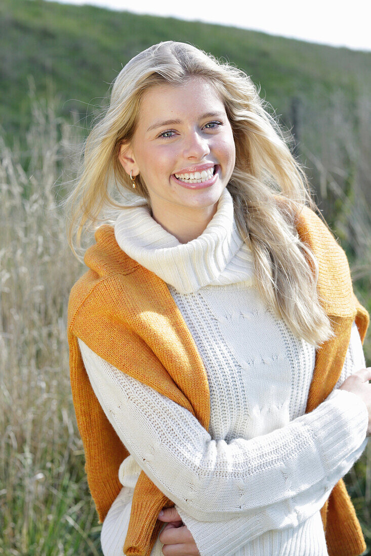 Junge blonde Frau in weißem Rollkragenpullover, mit gelbem Strickpulli über den Schultern in der Natur