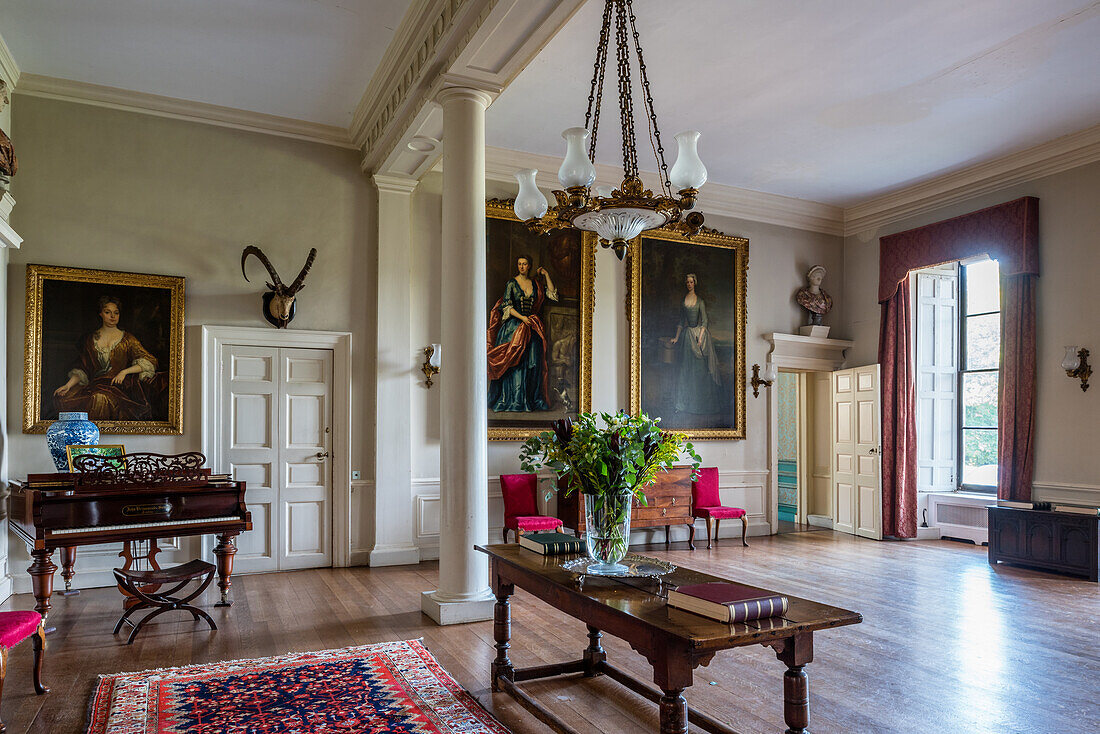 Table, piano, and large gold-frame paintings in the reception room of an English country house