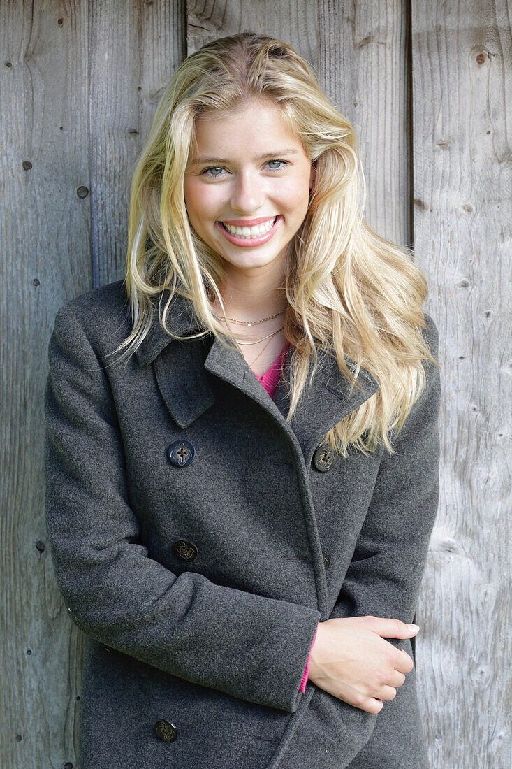 Young blonde woman in a gray wool coat in front of a wooden wall