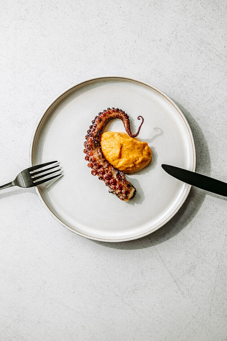 Grilled octopus tentacle, sweet potato puree, black cutlery, on a white plate, white background