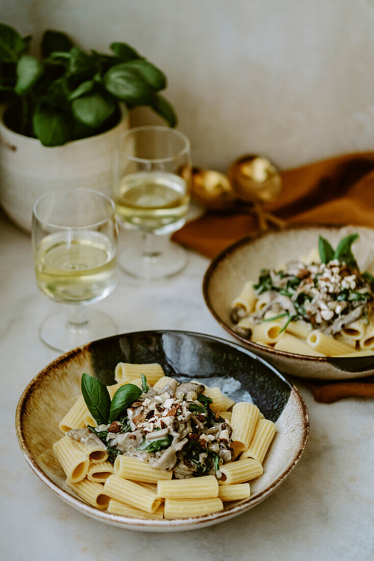 Rigatoni pasta dish, with cream, mushroom, spinach, almond and grated parmesan, in handmade plates, and glass of white wine, on a marble table, basil plant in the background