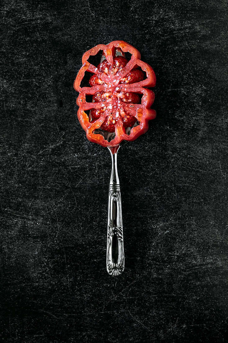 Slice of beef heart tomato, placed on a silver pie server, on a black textured background