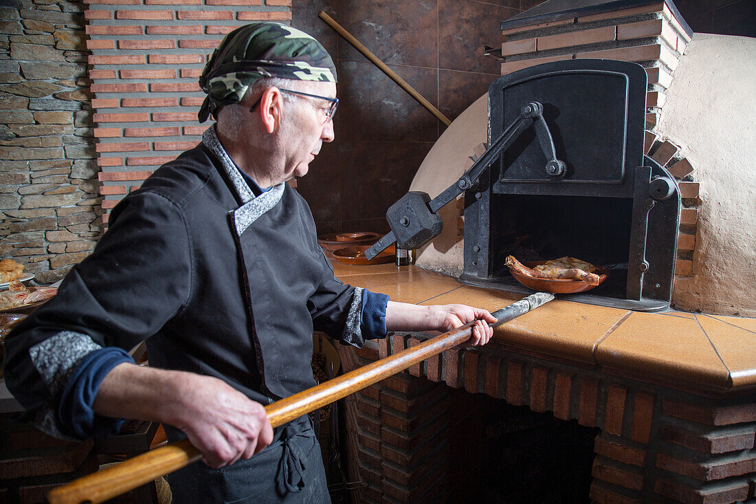Professional male cook in uniform putting bowl with meat in wood burning oven