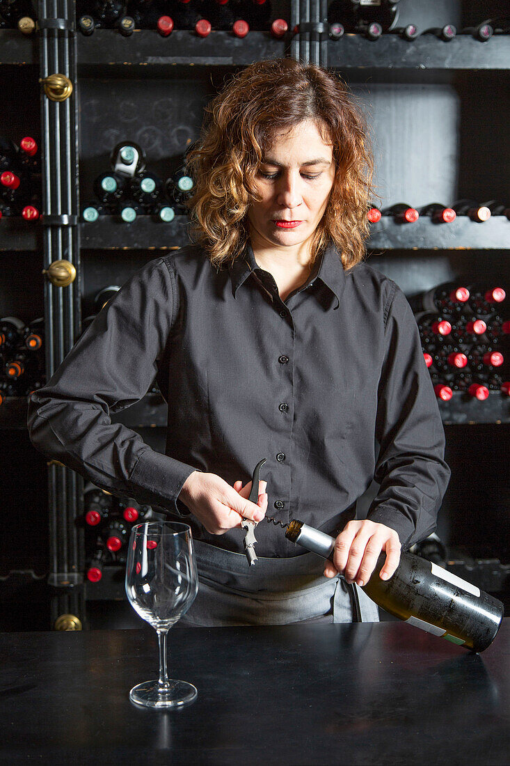 Focused female barkeeper in apron opening glass bottle of wine while standing at counter with wineglass during work in bar