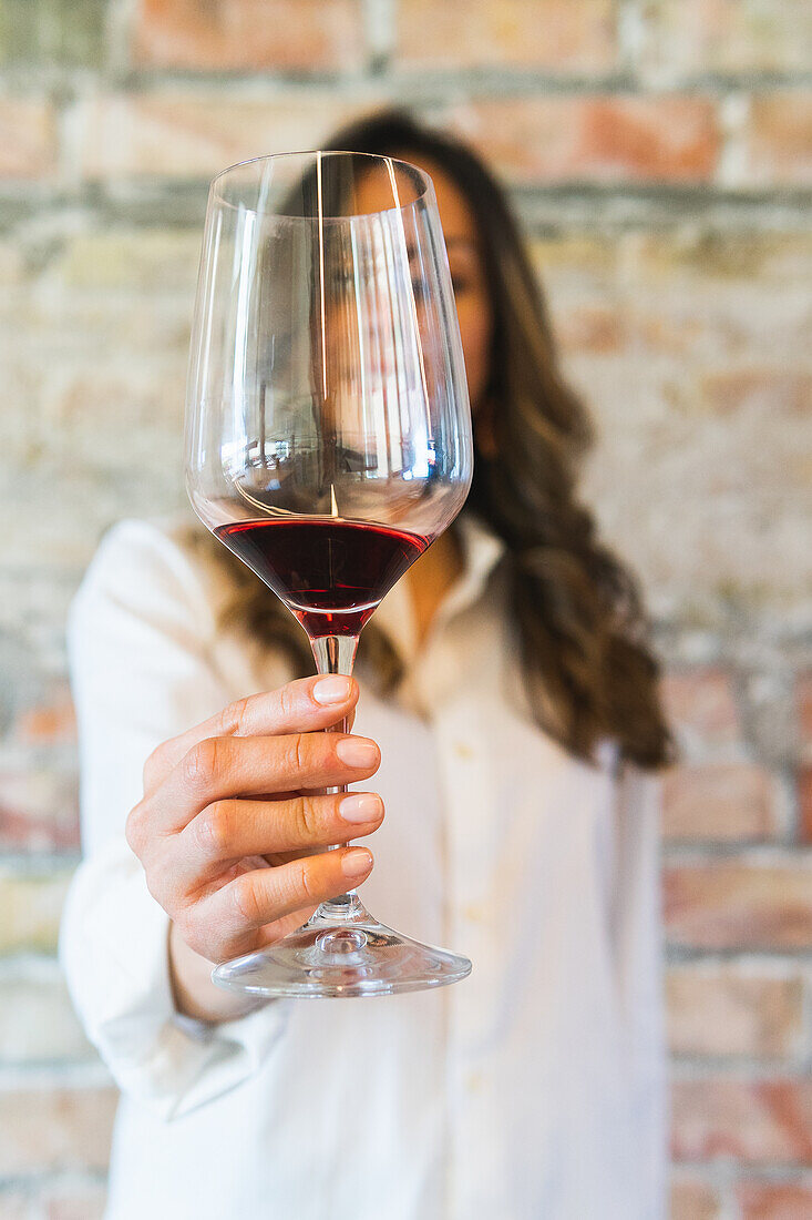 Soft focus of unrecognizable female sommelier in white shirt demonstrating glass goblet of red wine against brick wall in restaurant