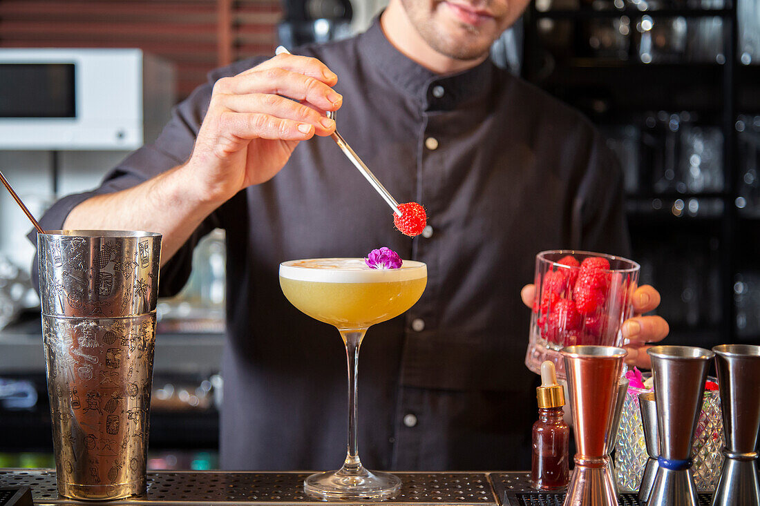 Crop unrecognizable male bartender in black shirt decorating fresh sour alcoholic cocktail with petals and raspberries while standing at counter with various tools in contemporary bar